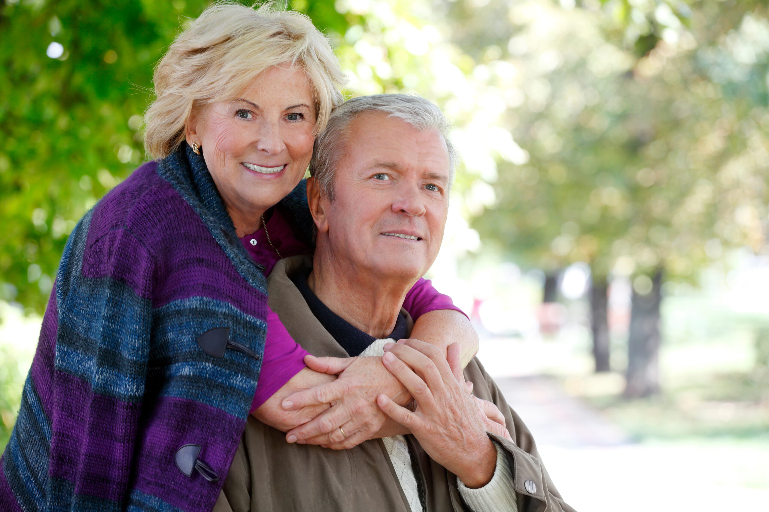 Close-up outdoor portrait of a senior couple smiling to the camera while elderly woman is hugging old man
